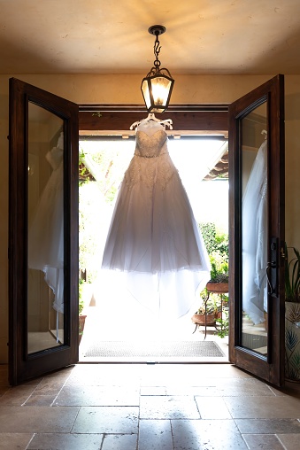 An elegant wedding dress hangs from an ornate chandelier suspended in a room with large windows
