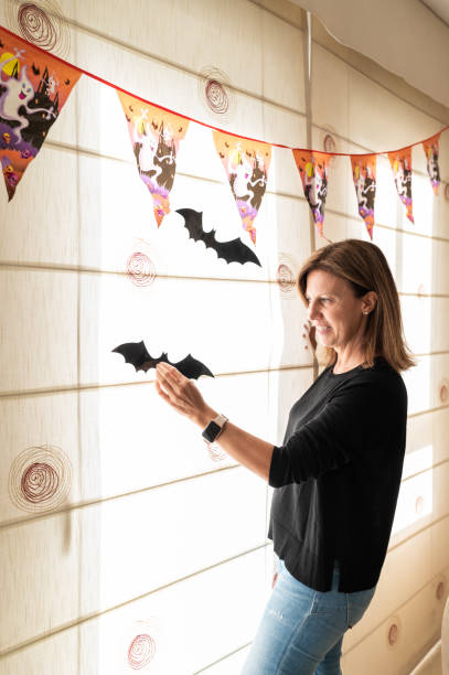 Vertical photo of a mature woman decorating home for Halloween with bat from paper and garlands