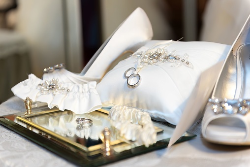 A closeup of wedding accessories featuring two gold rings, a white bridal bouquet, and a small ring bearer pillow