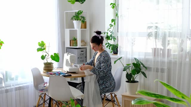 A woman sews tulle on an electric sewing machine in a white modern interior of a house with large windows, house plants. Comfort in the house, a housewife's hobby
