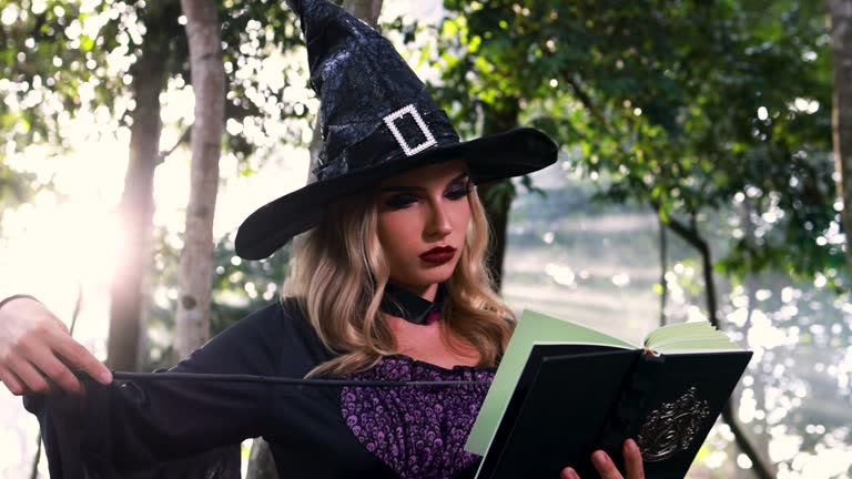 The beautiful woman in a witch  Halloween costume conjures, holds a magic book and a magic sparkle wand in her hand, and reads spell white magic smoke rises from a boiling jar.