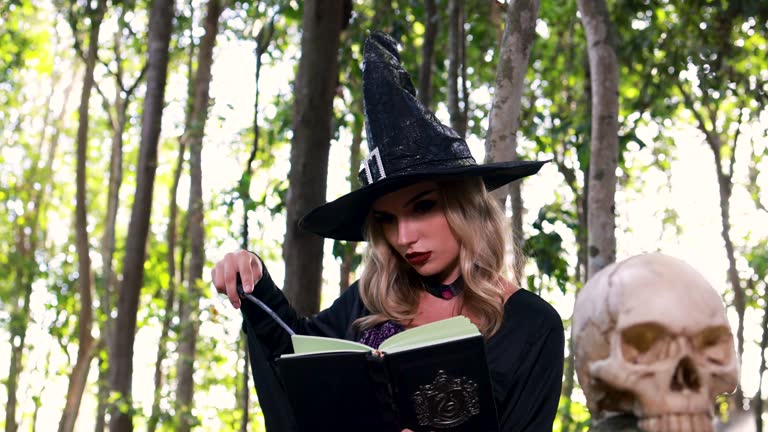 The beautiful woman in a witch  Halloween costume conjures, holds a magic book and a magic sparkle wand in her hand, and reads spell white magic smoke rises from a boiling jar.