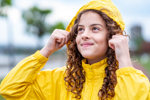 A teenage girl walking by the river has put on a raincoat because it is raining.
