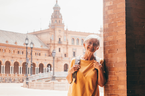 Happy senior woman tourist in yellow visiting Plaza de Espana in Seville. Traveler with backpack and large smile enjoying freedom and pleasure to travel
