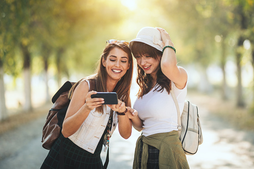 Two beautiful young smiling women with backpacks on their back are looking at the photos on the smartphone during walking along the autumn sunny avenue.