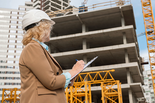 Senior woman entrepreneur makes notes on clipboard of building processes at construction site. Concept of working moments and production low angle shot