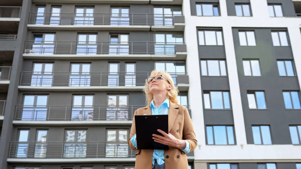 Senior woman entrepreneur looks around making notes on clipboard on apartment building background. Concept of working as real estate agent low angle shot