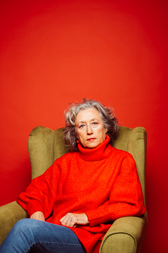 Studio portrait of a senior woman wearing blue jeans red sweater, sitting on a green armchair,  in front a red background