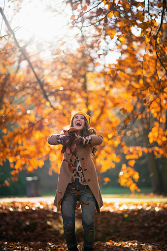 Hello september. happy trendy woman in beige coat and orange hat throws up autumn leaves outdoors in the city park in autumn.