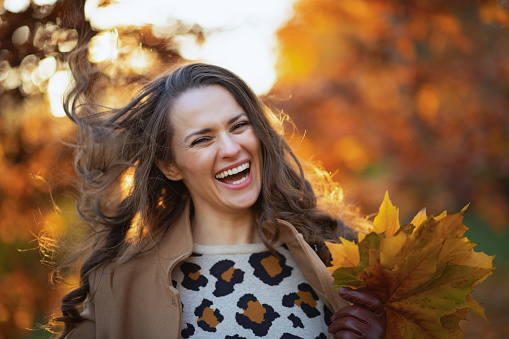 Hello november. smiling young female in beige coat with autumn yellow leaves outdoors in the city park in autumn.