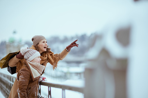 happy stylish mother and child in coat, hat, scarf and mittens with shopping bags pointing at something outdoors in the city in winter.