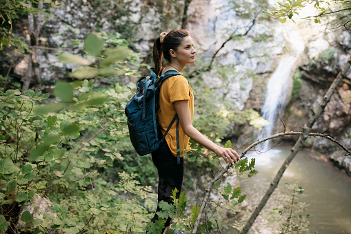 Pretty female with backpack looking mountain waterfall while hiking in forest. Woman tourist standing against waterfall from mountain on hiking trip.