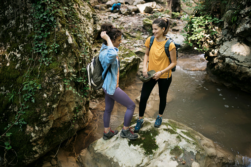 Young female friends standing and talking by mountain stream while hiking in forest. Two women taking break while hiking on forest trail.