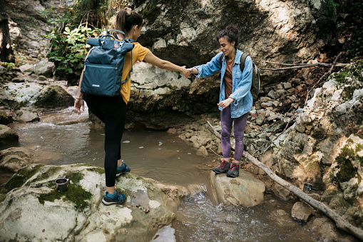 Young woman helping female friend giving her hand to cross a mountain creek while hiking. Two female hiker crossing mountain stream together.