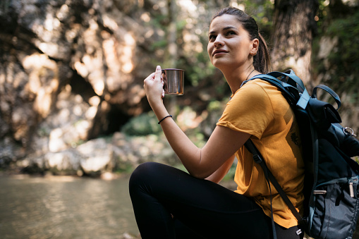 Beautiful young woman taking a coffee break while hiking in forest. Young female hiker sitting by mountain stream holding coffee mug and looking away.