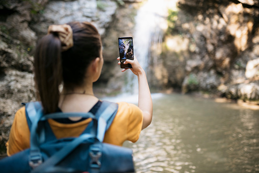 Rear view of a young woman hiker taking photos of a waterfall with her mobile phone. Female on hiking trip taking pictures of beautiful waterfall using her cell phone.