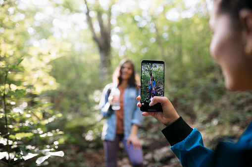 Close-up of a woman taking photos of her friend in nature during a hiking trip. Female friends taking photos on forest hike with focus on mobile phone screen.