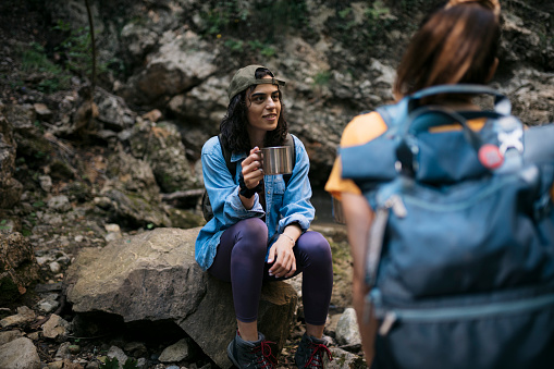 Two young women taking a break from hiking and drinking coffee. Female friends sitting on rocks drinking coffee and talking while taking a break from hiking on forest trail.