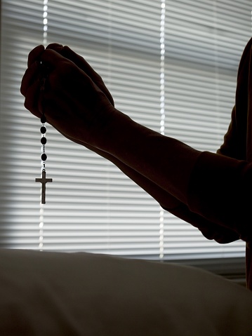 Hands in prayer with Rosary beads in silhouette. Darkened room with arms and hands shown in prayer in front of window blinds. Crucifix dangling on string of beads below praying hands with copy space.