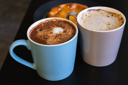 Two Cappuccino cups. Mugs full of coffee with milk foam and cinnamon powder stand on a black table, close up photo with selective soft focus