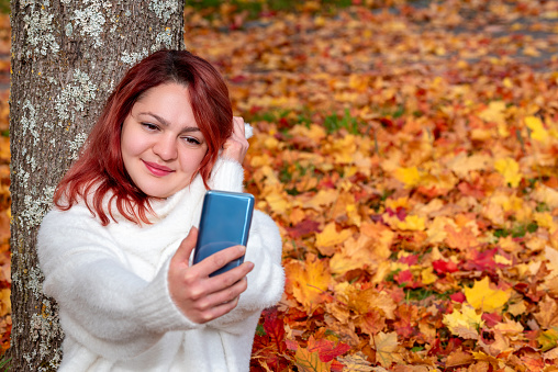 Autumn scenery with a beautiful woman taking selfies with her phone. Woman wearing a white sweater against a background of autumn leaves