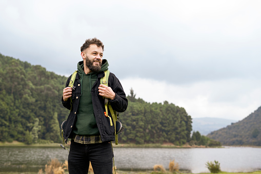 Young man with beard standing looking at the landscape, with the lake and mountains in the background, carrying a backpack
