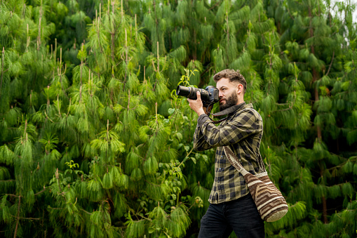 Young man with a beard and a plaid shirt, holds his camera to photograph the landscape, the mountains, trees and lake on a day of hiking through nature