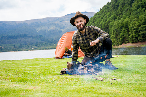 Young man with a beard, hat and plaid shirt sitting on the grass with the lake and mountains in the background, on a camping day lighting a bonfire to share with his friends