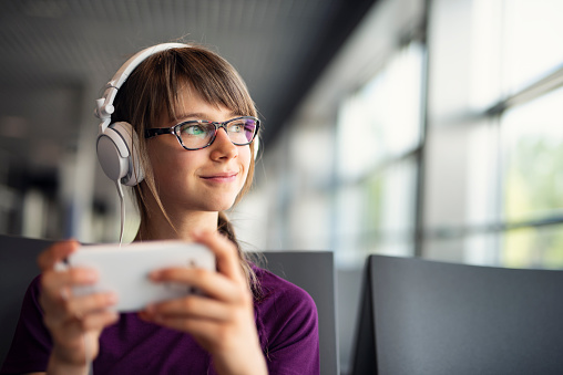 Little girl waiting at the airport. The girl is wearing headphones and playing with smartphone. \nNikon D810