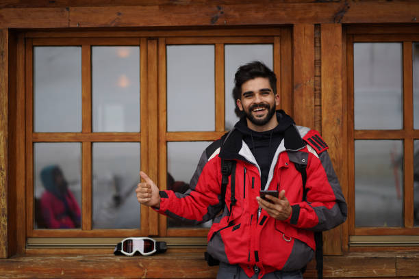 Happy handsome man wearing ski clothes standing outdoors holding a mobile phone looking at camera Happy handsome man wearing ski clothes standing outdoors in winter season, holding a mobile phone looking at camera, feeling confident and showing thumbs up. thumbs up for skiing stock pictures, royalty-free photos & images