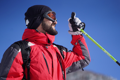 Cheerful man wearing winter jacket, cap and gloves, smiling and holding a ski pole on a clear sunny weather outdoors.