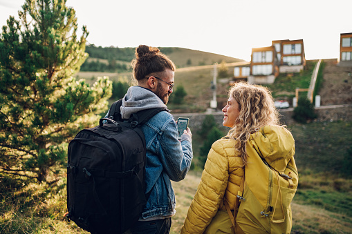 Rear view of a couple of travelers hiking on the beautiful mountain trail at warm sunny evening while using a map on a smartphone. Happy young man and woman on vacation in countryside. Copy space.