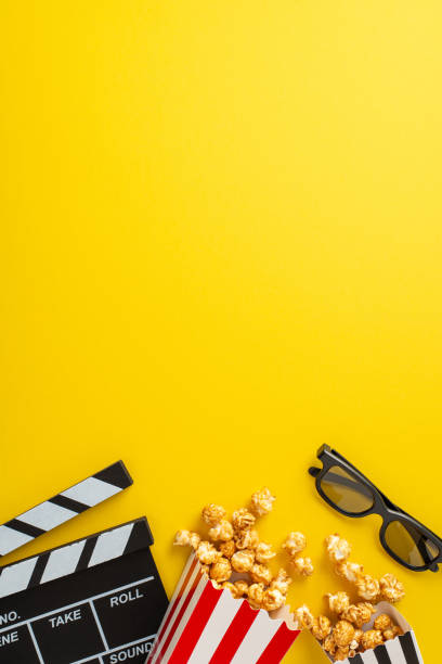 istock Cinematograph concept visualized: Vertical top view of striped containers, holding popcorn, 3D glasses, and a clapperboard. Presented on yellow canvas with vacant space for text or promotional content 1659934563