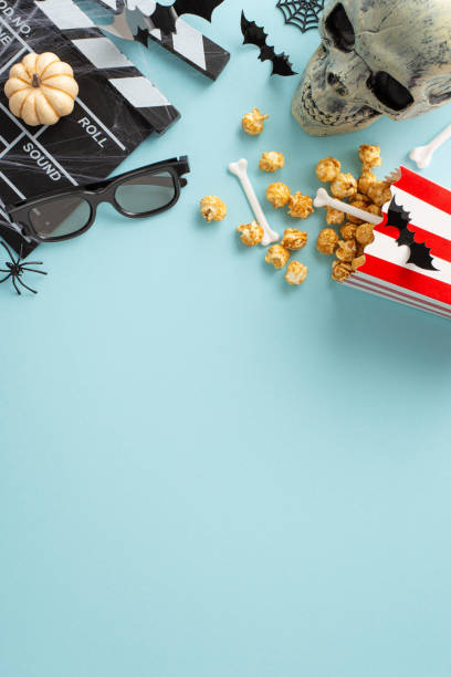istock Halloween movie magic. Vertical top view showcases thematic decor: popcorn box with bones, eerie insects, skull, pumpkin, spiderweb, bats, clapper, 3D glasses. Pastel blue backdrop has space for text 1659934780