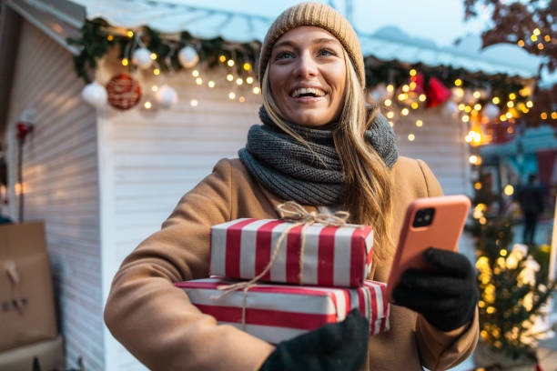 Excited young woman holding Christmas presents and using phone, on a Christmas market