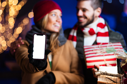 Young man and woman outdoors, celebrating Christmas. Woman is holding a phone with a white screen on.