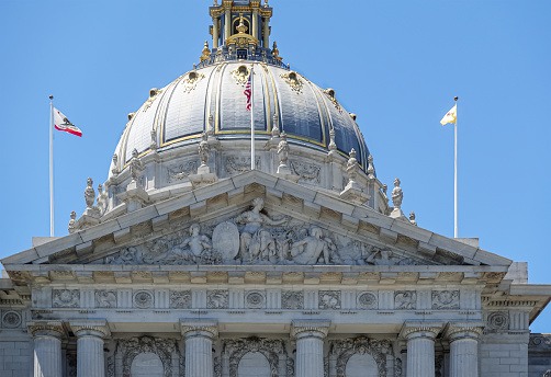 San Francisco, CA, USA - July 12, 2023: Gray stone City Hall east facade pediment above entrance with giant dome under blue sky. Golden decorations.  3 Flags on top: country, state, city