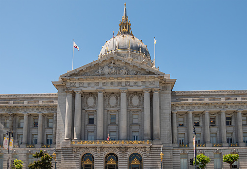 San Francisco, CA, USA - July 12, 2023: Gray stone City Hall east facade with pediment and giant dome under blue sky. Golden decorations at and on spire and above doors. 3 Flags on top.