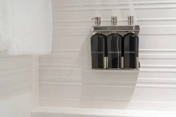 White hotel shower wall with bottles of body wash, liquid soap, hair conditioner, and shampoo in a chrome dispensers with a towel (Hotel amenities).