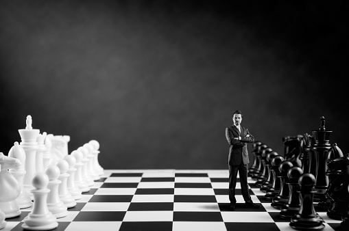 Businessman in suit with hands crossed is standing on a Chess Board near black Chess Pieces, processing for black and white look.