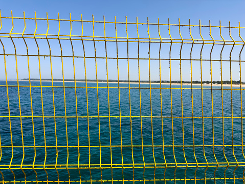 Blue sky behind a wire fence. Beach and sea in the background. Sunny summer day. Yellow wire mesh for security on the ferry pier. No people.