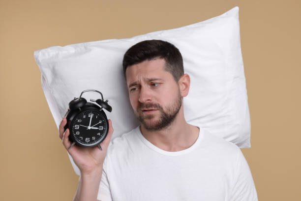 istock Sleepy man with pillow and alarm clock on beige background. Insomnia problem 1660041830