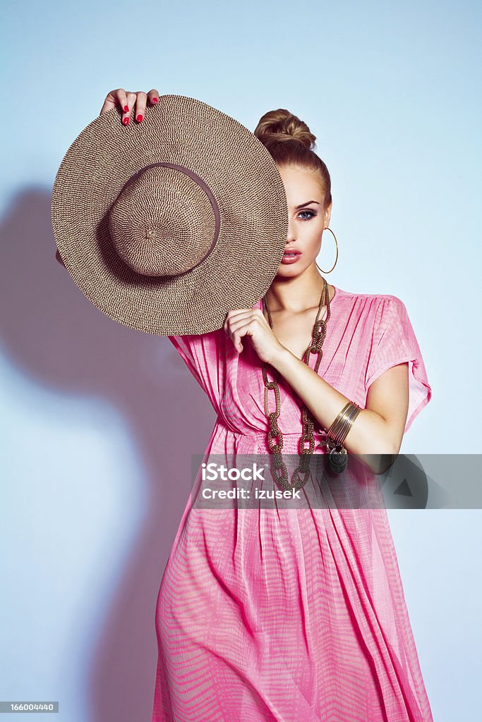 Glamour portrait of woman posing with sun hat Beautiful young blond woman wearing pink, summer dress and gold jewellery covering one eye with her sun hat. Studio shot, blue background. Summer portrait. Summer Stock Photo