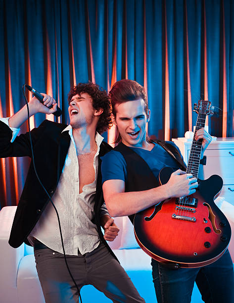 Young adult people having fun at the home party. Two guys pretending a music band, one of them singing into microphone, second playing a guitar.