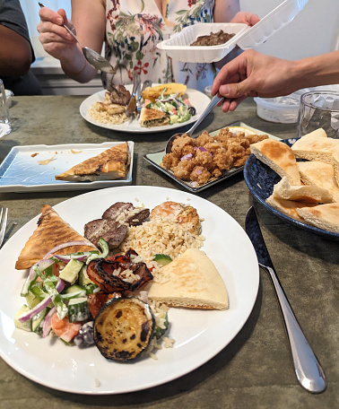 Members of a multi-racial family select from a variety of Greek takeout food at a summer dinner party in Metro Vancouver. The spanakopita, calamari and pita are set on Japanese serving plates.