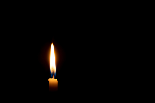 Single burning candle flame or light is glowing on a small yellow candle on black or dark background on table in church for Christmas, funeral or memorial service with copy space.