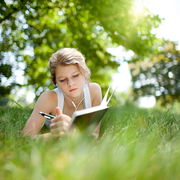 A young girl listening to music and studying in the park Beautiful teenage girl laying on the grass, writing in her notebook, listening to music 13 14 years child teenage girls lying down stock pictures, royalty-free photos & images