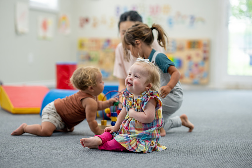 A sweet little girl with Down Syndrome plays on the floor at daycare with a few little friends and her Childcare provider.  She is dressed casually and has a smile on her face as she giggles joyfully.