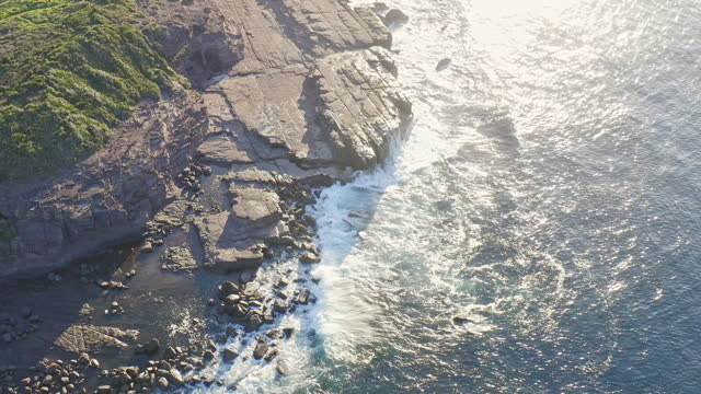 Wide aerial shot of ocean waves crashing over rocks on a sunny morning