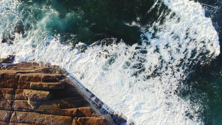 Aerial shot of ocean waves crashing against rocks on a sunny morning